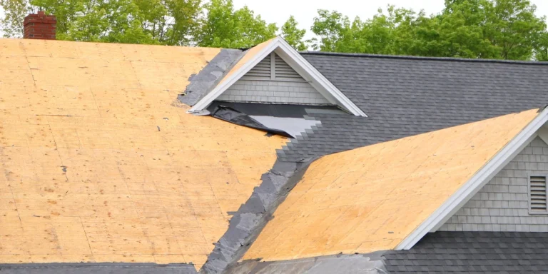 A close-up view of a residential roof replacement showing exposed plywood decking, black underlayment, and new architectural shingles being installed around a dormer.